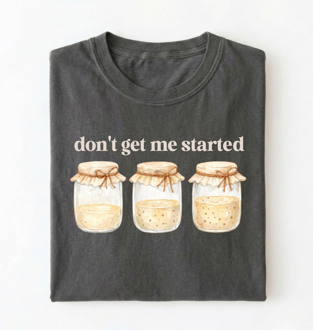 Flat lay of a pepper t-shirt featuring illustrated jars and text reading “Don’t Get Me Started. Funny sourdough starter perfect gift for bakers fermentation lovers and bread enthusiasts on a white background
