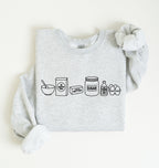 A light heather gray sweatshirt with a black line art graphic of baking ingredients (a bowl, flour bag, butter stick, sugar jar, vanilla bottle, and eggs) on the front. The sweatshirt is styled flat on a cream background with a gray scrunchie.