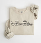 A beige crewneck sweatshirt with a black line art graphic of baking ingredients (bowl, flour bag, butter, sugar jar, vanilla, eggs) on the front, styled flat on a white background with a matching fabric scrunchie.
