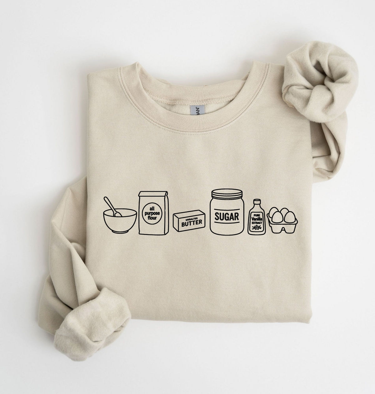 A beige crewneck sweatshirt with a black line art graphic of baking ingredients (bowl, flour bag, butter, sugar jar, vanilla, eggs) on the front, styled flat on a white background with a matching fabric scrunchie.