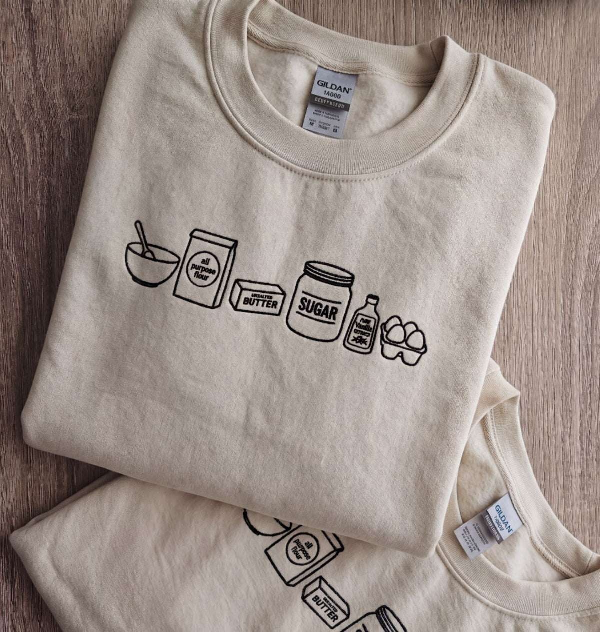 A folded cream-colored Gildan t-shirt with a black line art graphic of baking ingredients (bowl, flour, butter, sugar, syrup, eggs) on a wooden surface, with another t-shirt partially visible underneath.