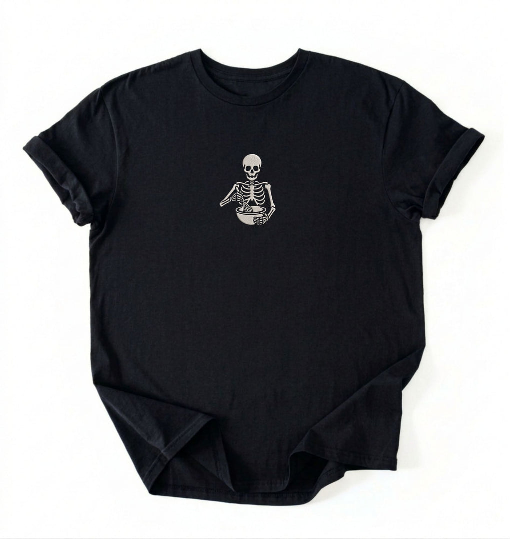 Flat lay of a a Black Comfort Colors shirt featuring an embroidered skeleton whisking ingredients in a mixing bowl on a white background 