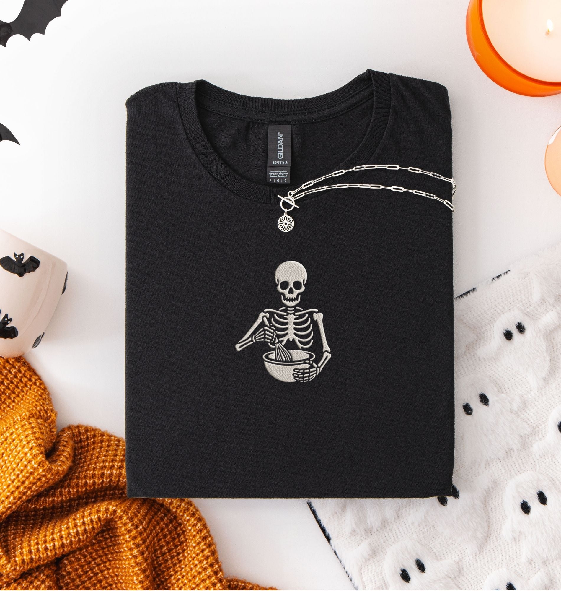 Flat lay of a a black Comfort Colors shirt featuring an embroidered skeleton whisking ingredients in a mixing bowl on a white background 