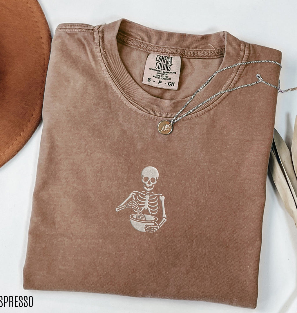 Flat lay of a a brown Comfort Colors shirt featuring an embroidered skeleton whisking ingredients in a mixing bowl on a white background 