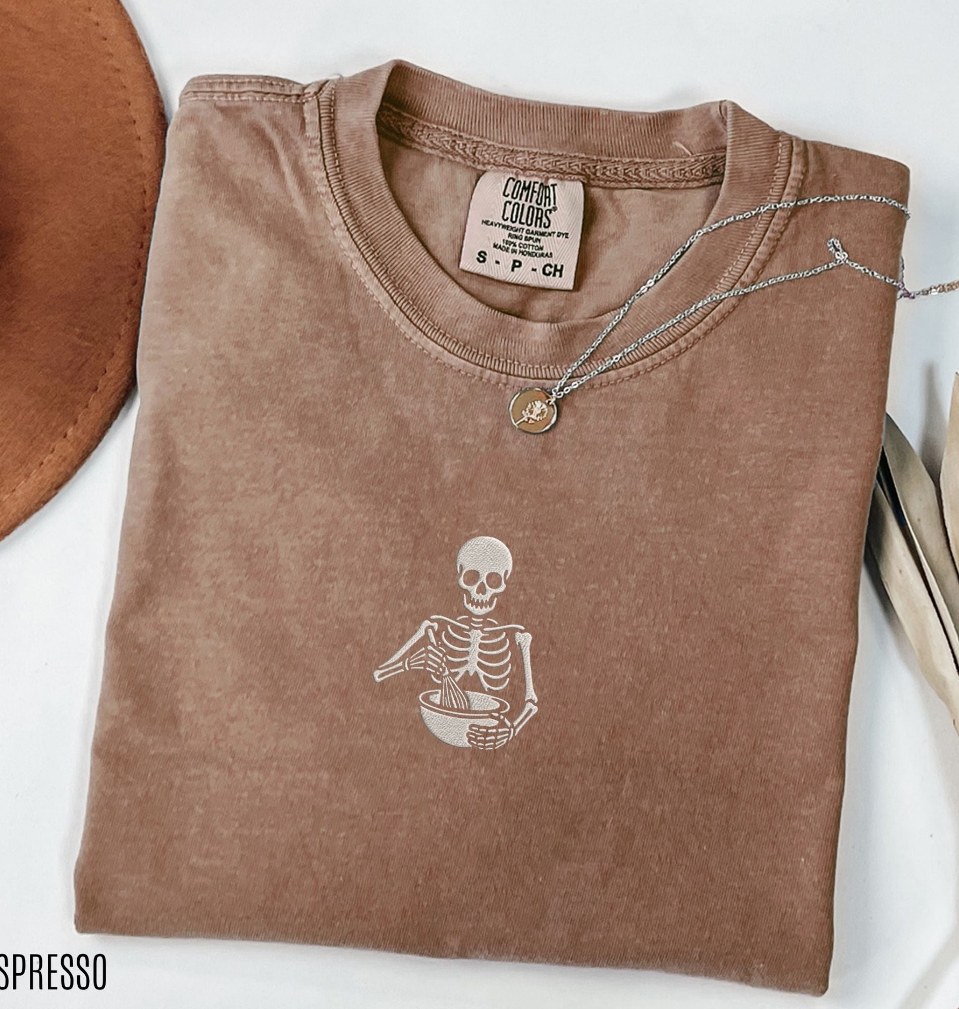 Flat lay of a a brown Comfort Colors shirt featuring an embroidered skeleton whisking ingredients in a mixing bowl on a white background 