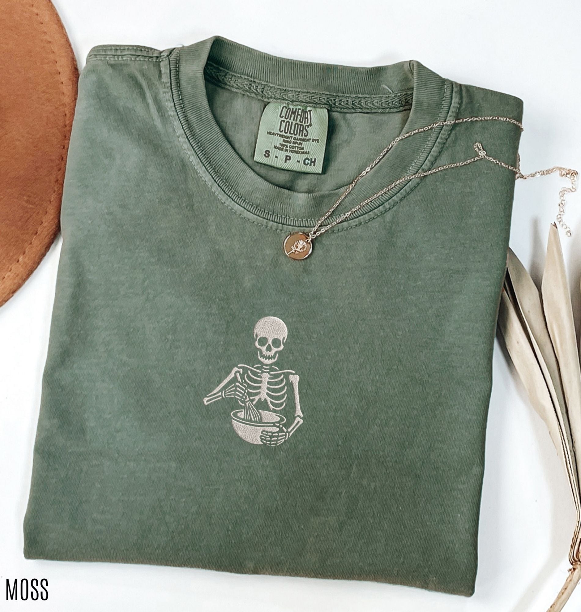 Flat lay of a a military green Comfort Colors shirt featuring an embroidered skeleton whisking ingredients in a mixing bowl on a white background 