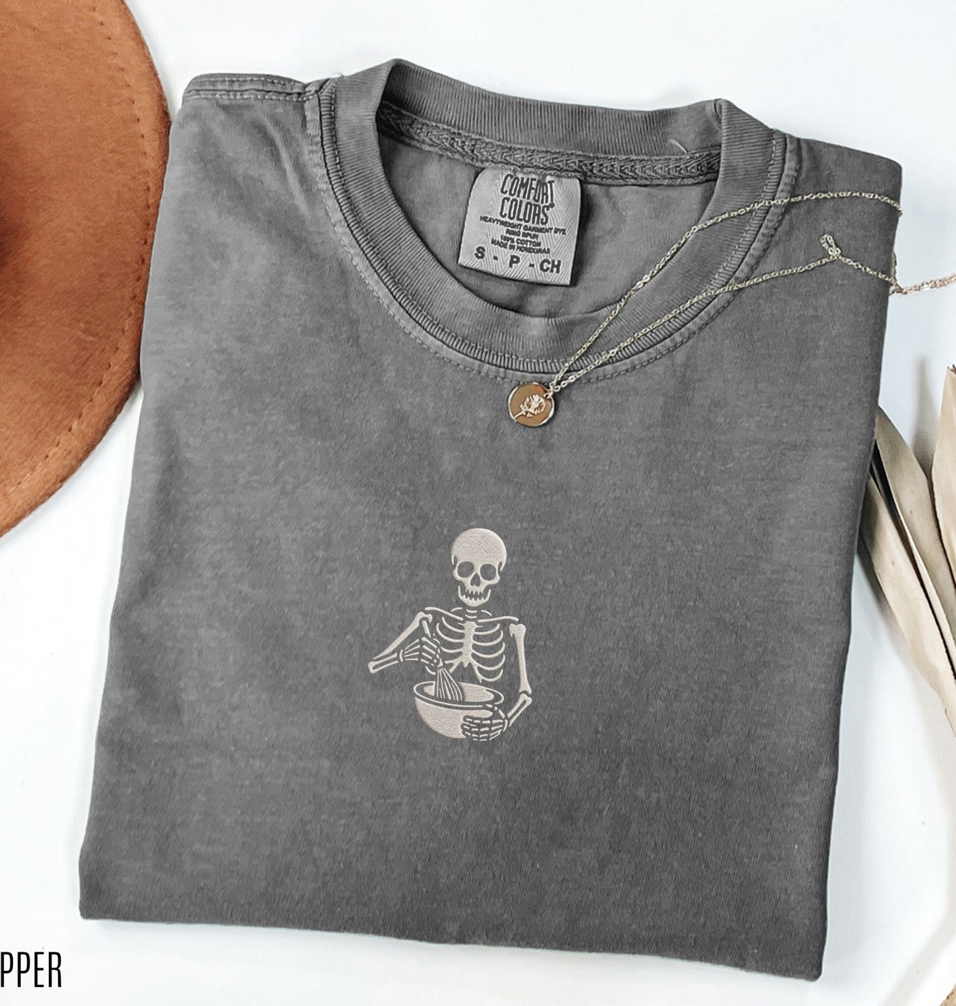 Flat lay of a a dark gray Comfort Colors shirt featuring an embroidered skeleton whisking ingredients in a mixing bowl on a white background 
