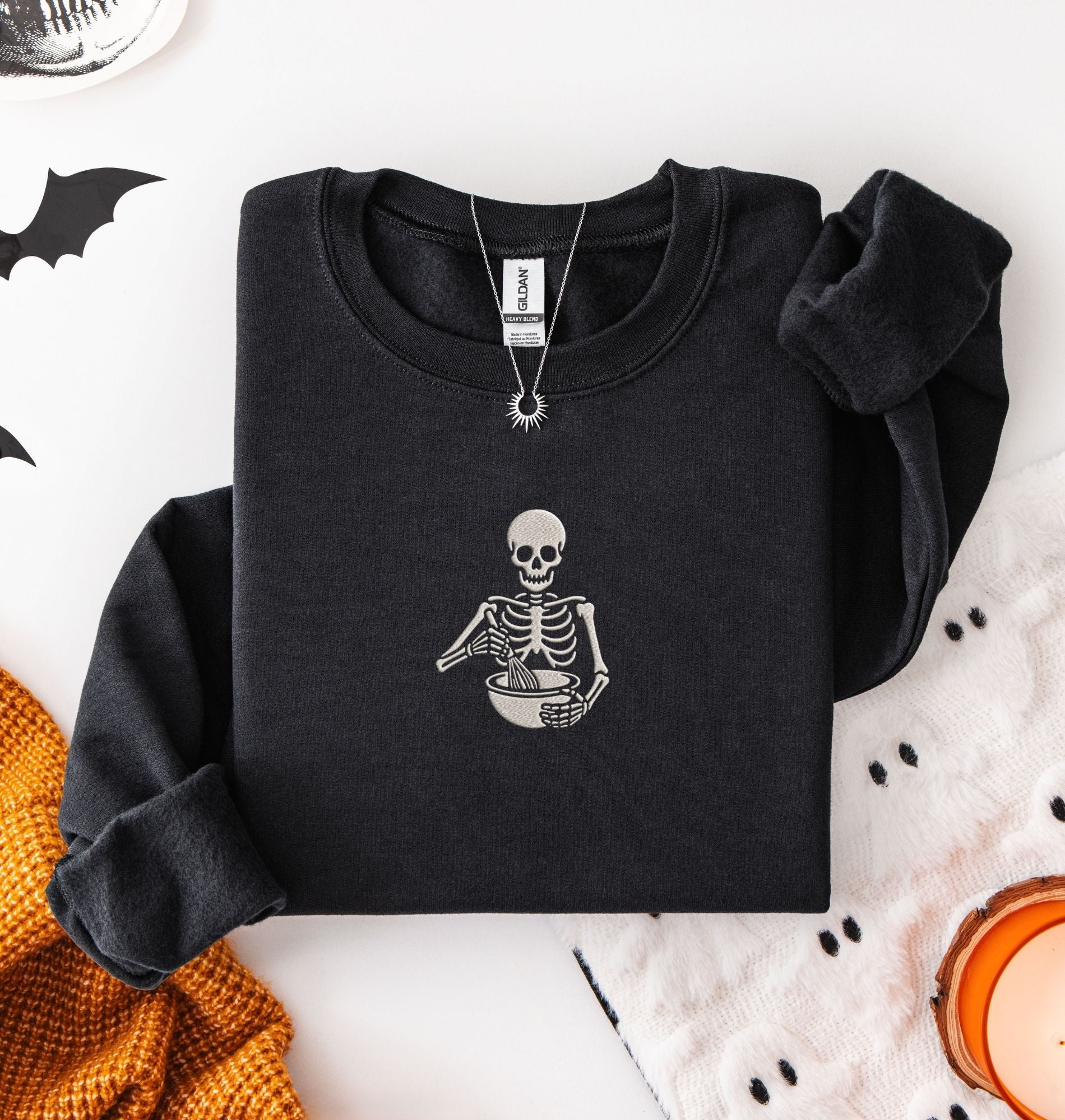 Flat lay of a Black Gildan sweatshirt featuring an embroidered skeleton whisking ingredients in a mixing bowl on a white background 
