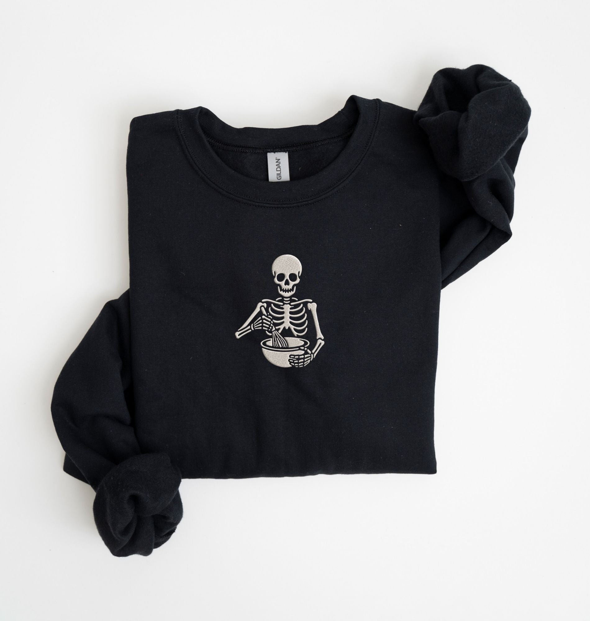 Flat lay of a Black Gildan sweatshirt featuring an embroidered skeleton whisking ingredients in a mixing bowl on a white background 
