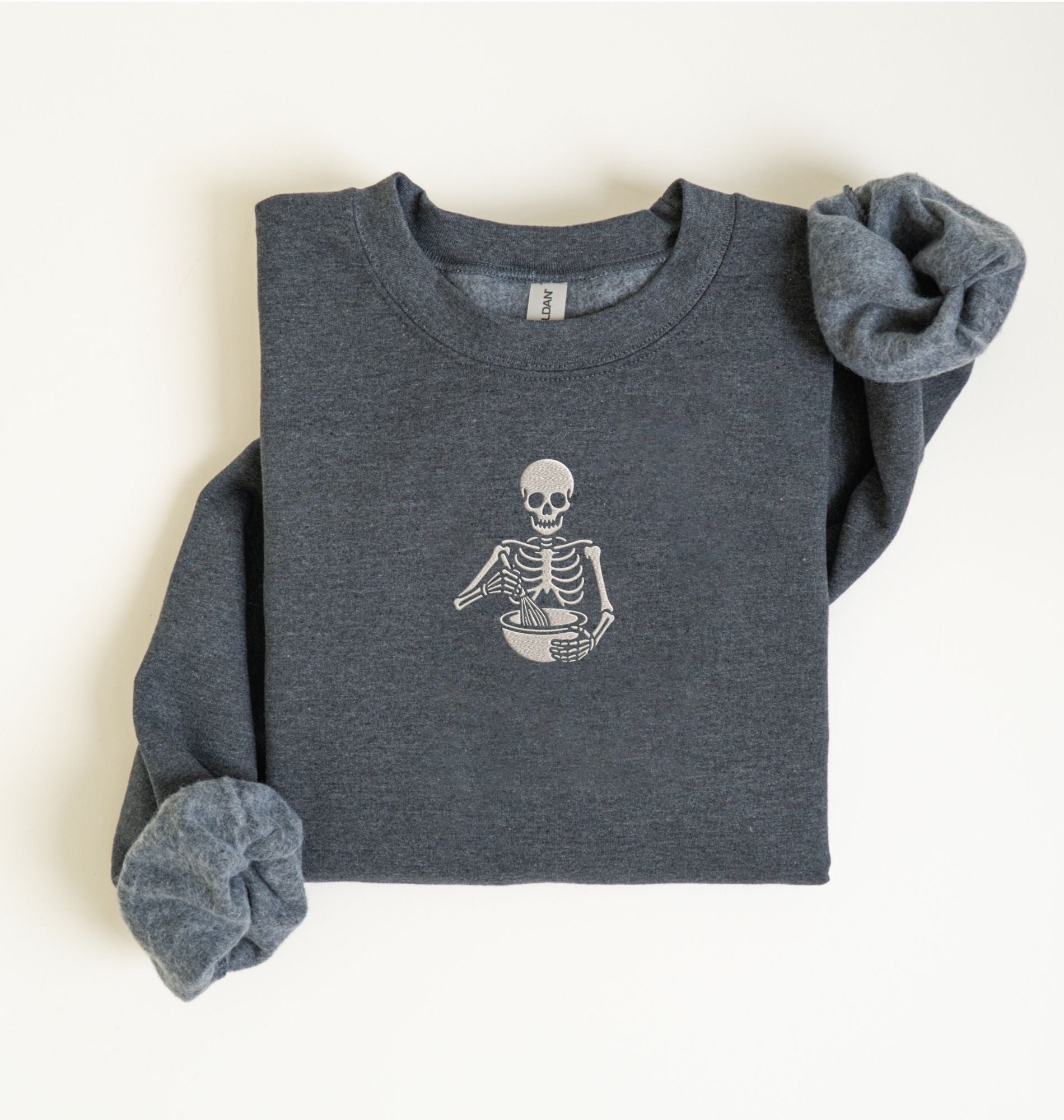 Flat lay of a dark heather gray Gildan sweatshirt featuring an embroidered skeleton whisking ingredients in a mixing bowl on a white background 