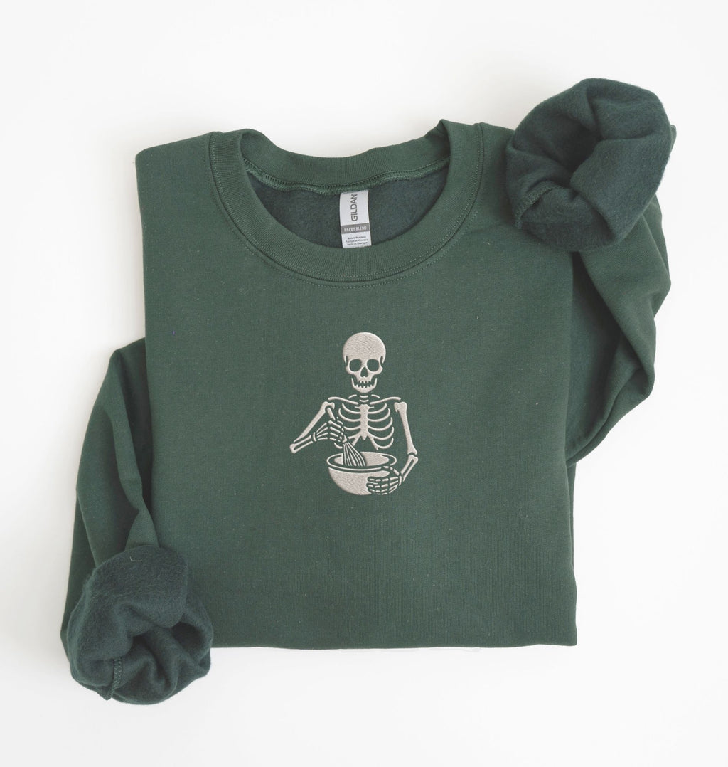 Flat lay of a forest green Gildan sweatshirt featuring an embroidered skeleton whisking ingredients in a mixing bowl on a white background 