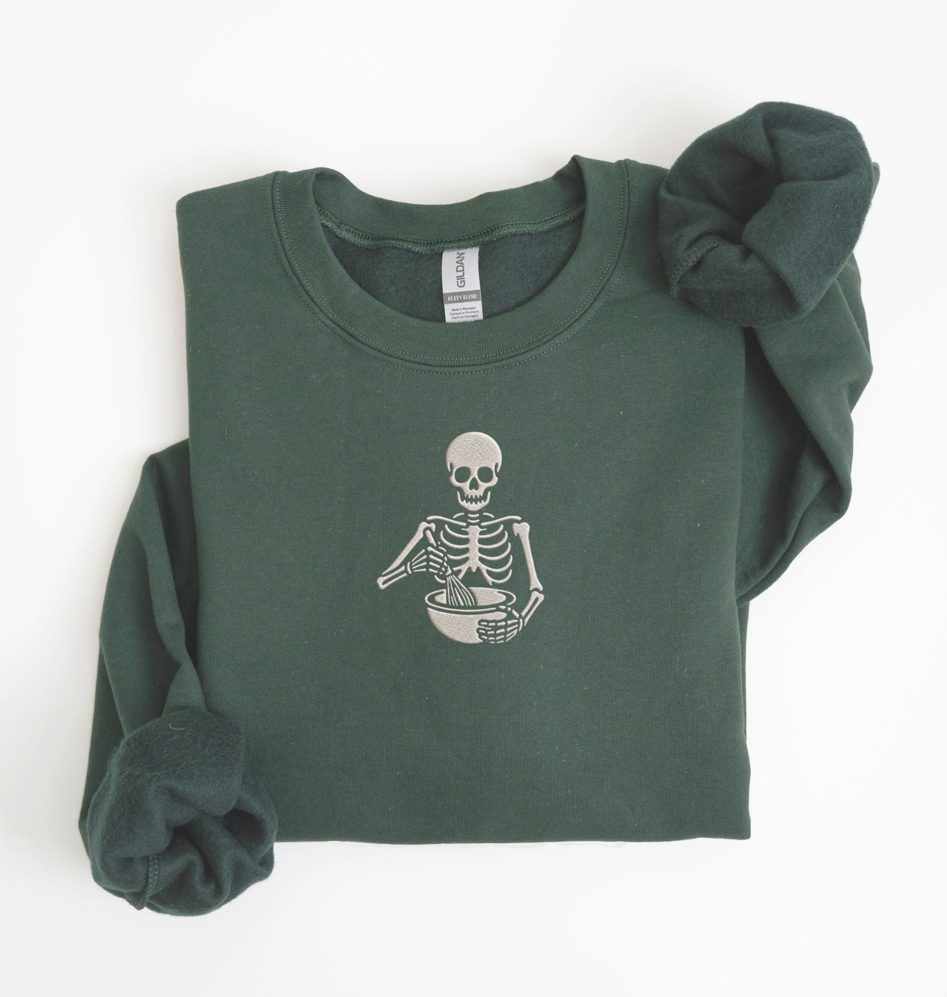 Flat lay of a forest green Gildan sweatshirt featuring an embroidered skeleton whisking ingredients in a mixing bowl on a white background 