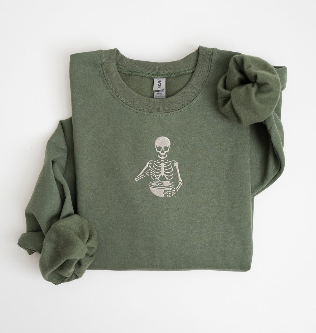 Flat lay of a military green Gildan sweatshirt featuring an embroidered skeleton whisking ingredients in a mixing bowl on a white background 