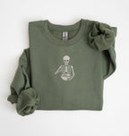 Flat lay of a military green Gildan sweatshirt featuring an embroidered skeleton whisking ingredients in a mixing bowl on a white background 