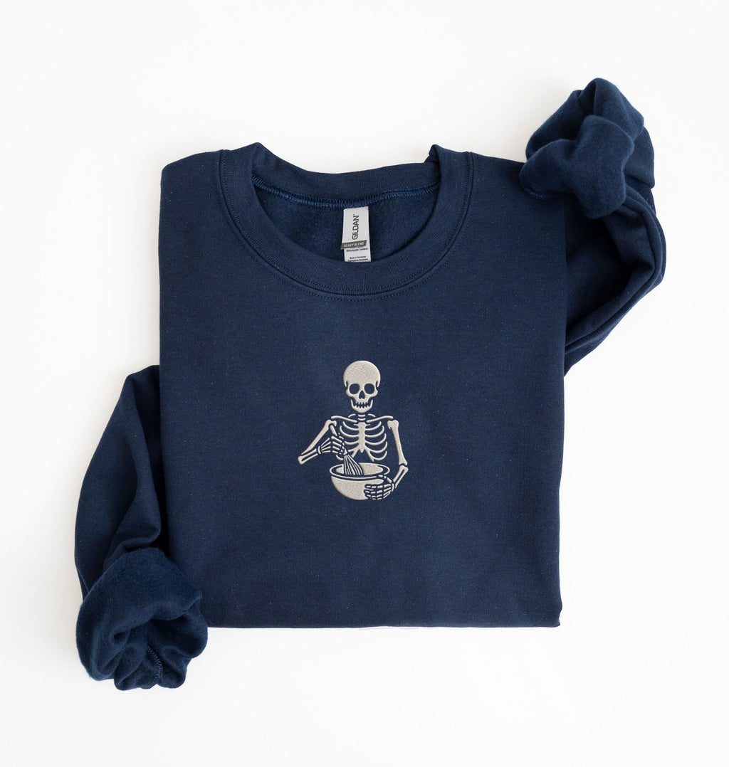 Flat lay of a navy blue Gildan sweatshirt featuring an embroidered skeleton whisking ingredients in a mixing bowl on a white background 