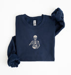 Flat lay of a navy blue Gildan sweatshirt featuring an embroidered skeleton whisking ingredients in a mixing bowl on a white background 