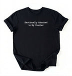 A folded black-colored t-shirt with the black printed text 'Emotionally Attached to My Starter' on the front, displayed against a clean white background.