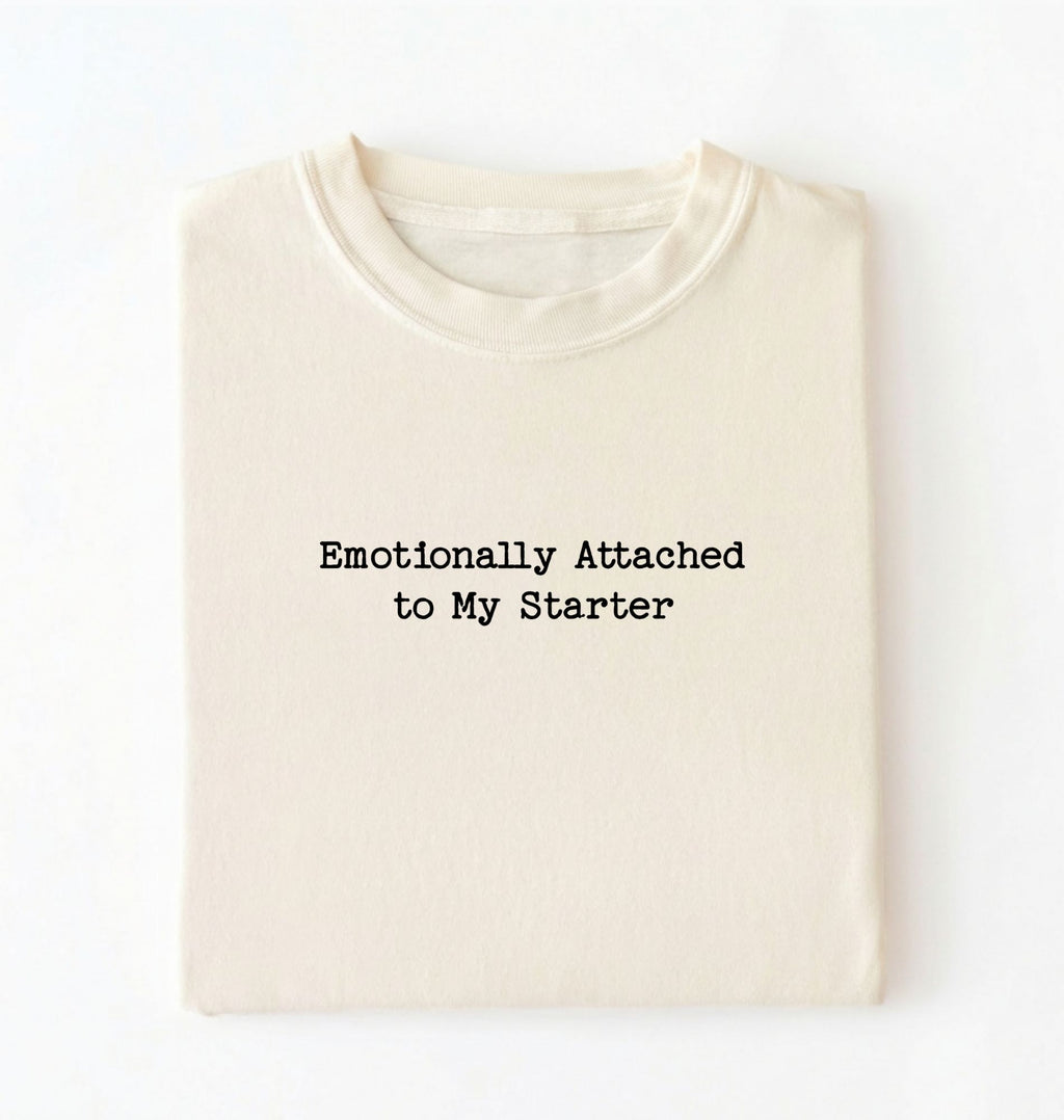 A folded cream-colored t-shirt with the black printed text 'Emotionally Attached to My Starter' on the front, displayed against a clean white background.