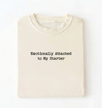 A folded cream-colored t-shirt with the black printed text 'Emotionally Attached to My Starter' on the front, displayed against a clean white background.