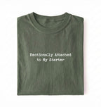 A folded olive green t-shirt with the text 'Emotionally Attached to My Starter' printed in white typewriter-style font on the front, set against a plain white background.