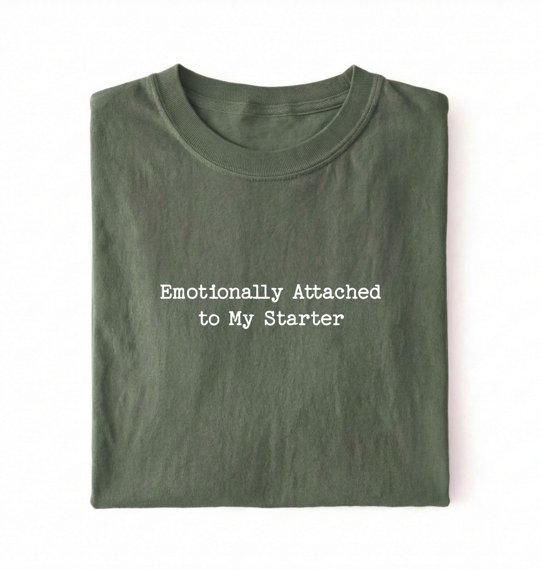 A folded olive green t-shirt with the text 'Emotionally Attached to My Starter' printed in white typewriter-style font on the front, set against a plain white background.