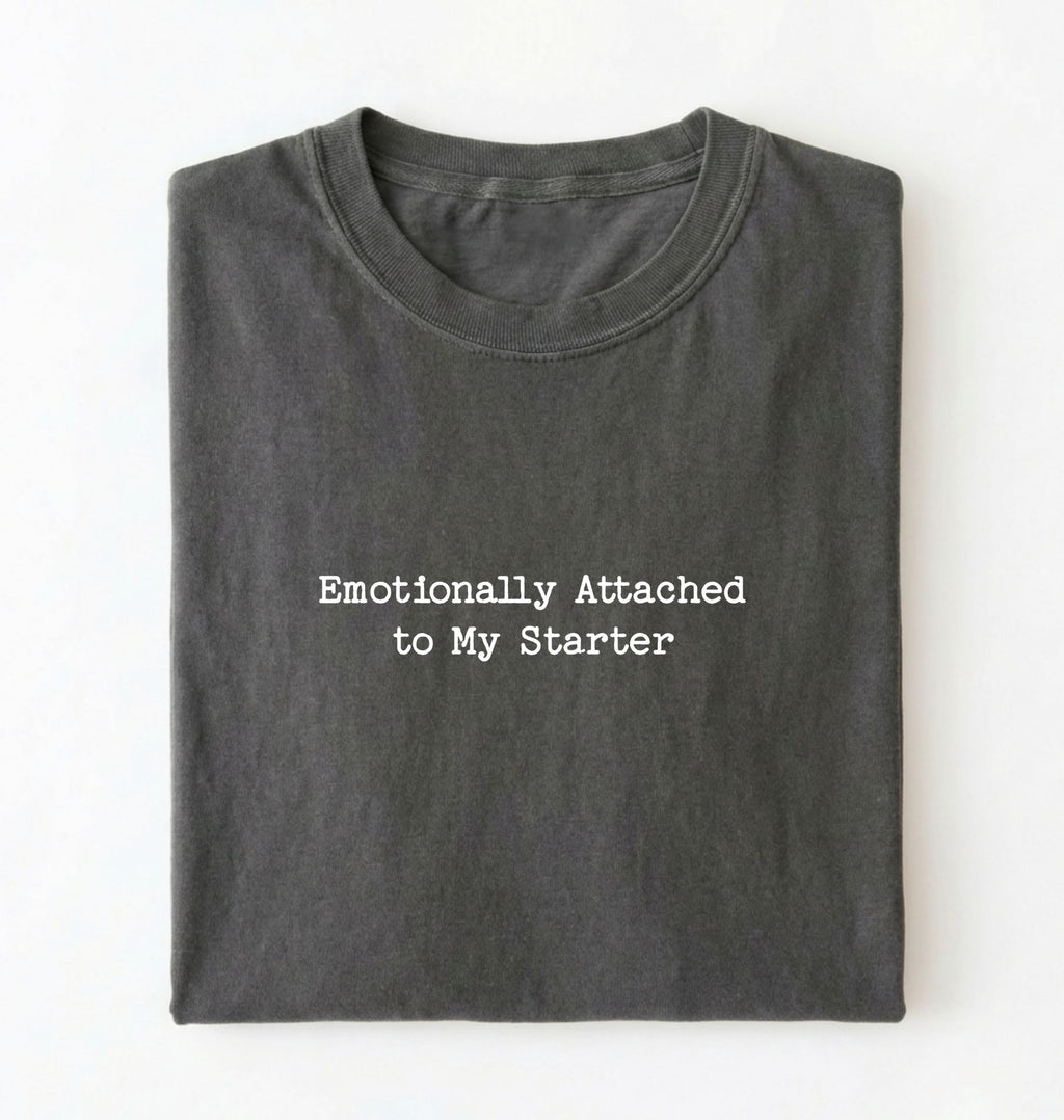 A folded olive pepper t-shirt with the text 'Emotionally Attached to My Starter' printed in white typewriter-style font on the front, set against a plain white background.