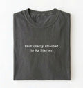 A folded olive pepper t-shirt with the text 'Emotionally Attached to My Starter' printed in white typewriter-style font on the front, set against a plain white background.