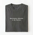 A folded olive pepper t-shirt with the text 'Emotionally Attached to My Starter' printed in white typewriter-style font on the front, set against a plain white background.