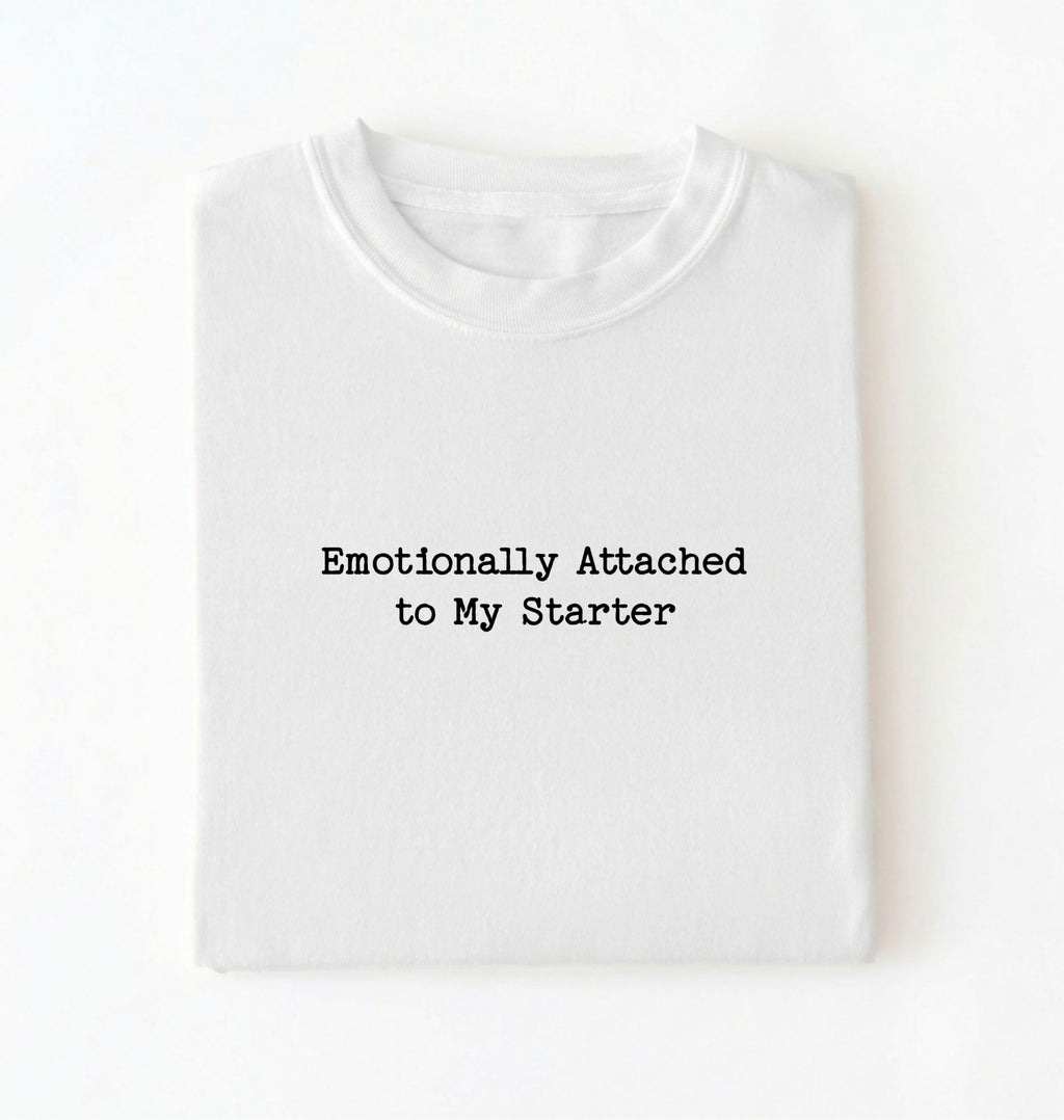 A folded white t-shirt with the text 'Emotionally Attached to My Starter' printed in a black typewriter-style font on the front, displayed against a clean white background.