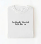 A folded white t-shirt with the text 'Emotionally Attached to My Starter' printed in a black typewriter-style font on the front, displayed against a clean white background.