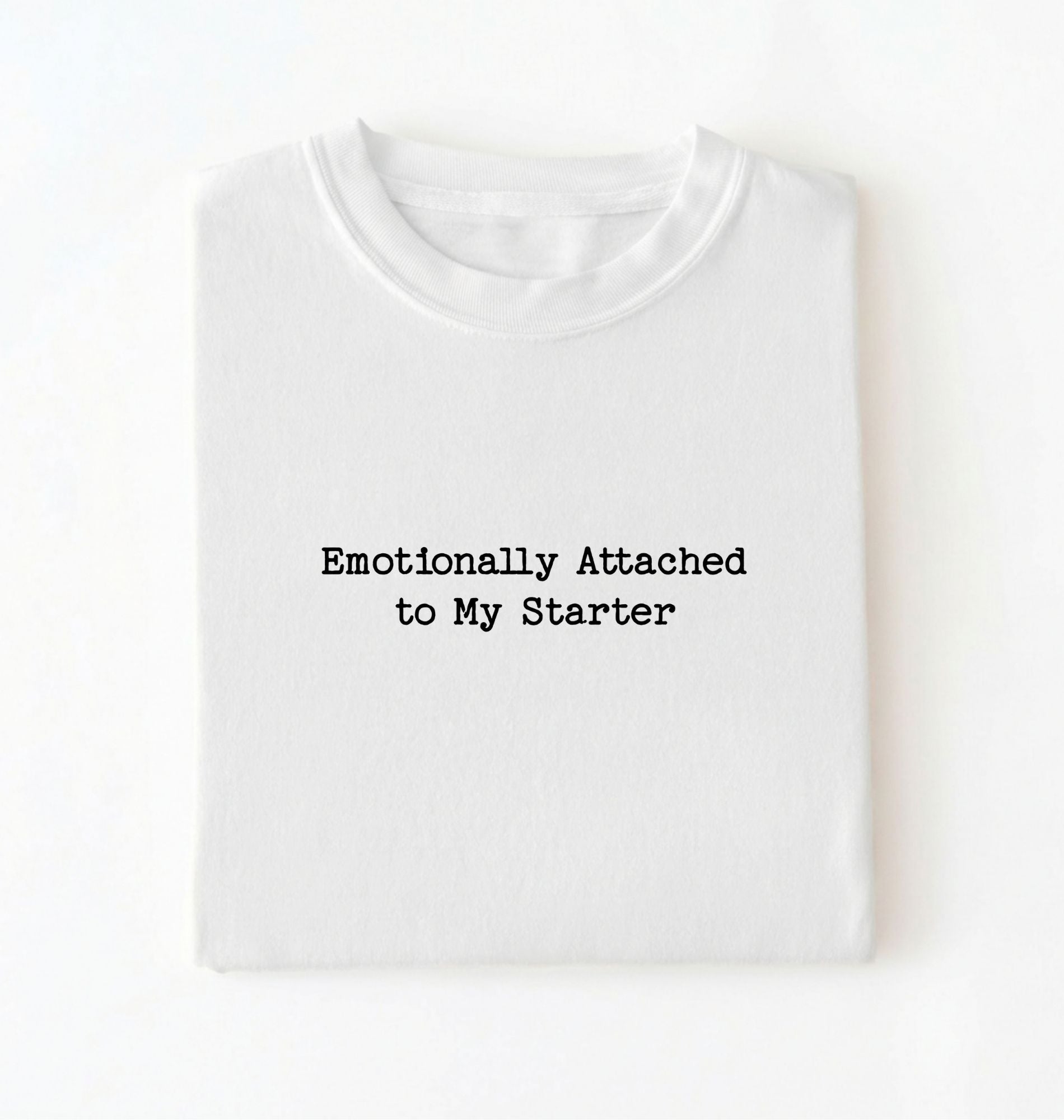 A folded white t-shirt with the text 'Emotionally Attached to My Starter' printed in a black typewriter-style font on the front, displayed against a clean white background.