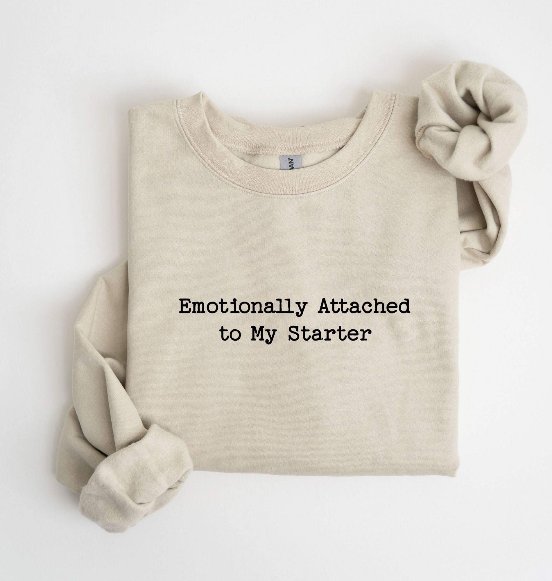A folded beige sweatshirt with the text 'Emotionally Attached to My Starter' printed in black on the front, styled with rolled sleeves on a white background.