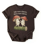 Flat lay of a dark_chocolate t-shirt with two dancing mushrooms and the phrase take more chances dance more dances, whimsical cottagecore gift tee on a white background 