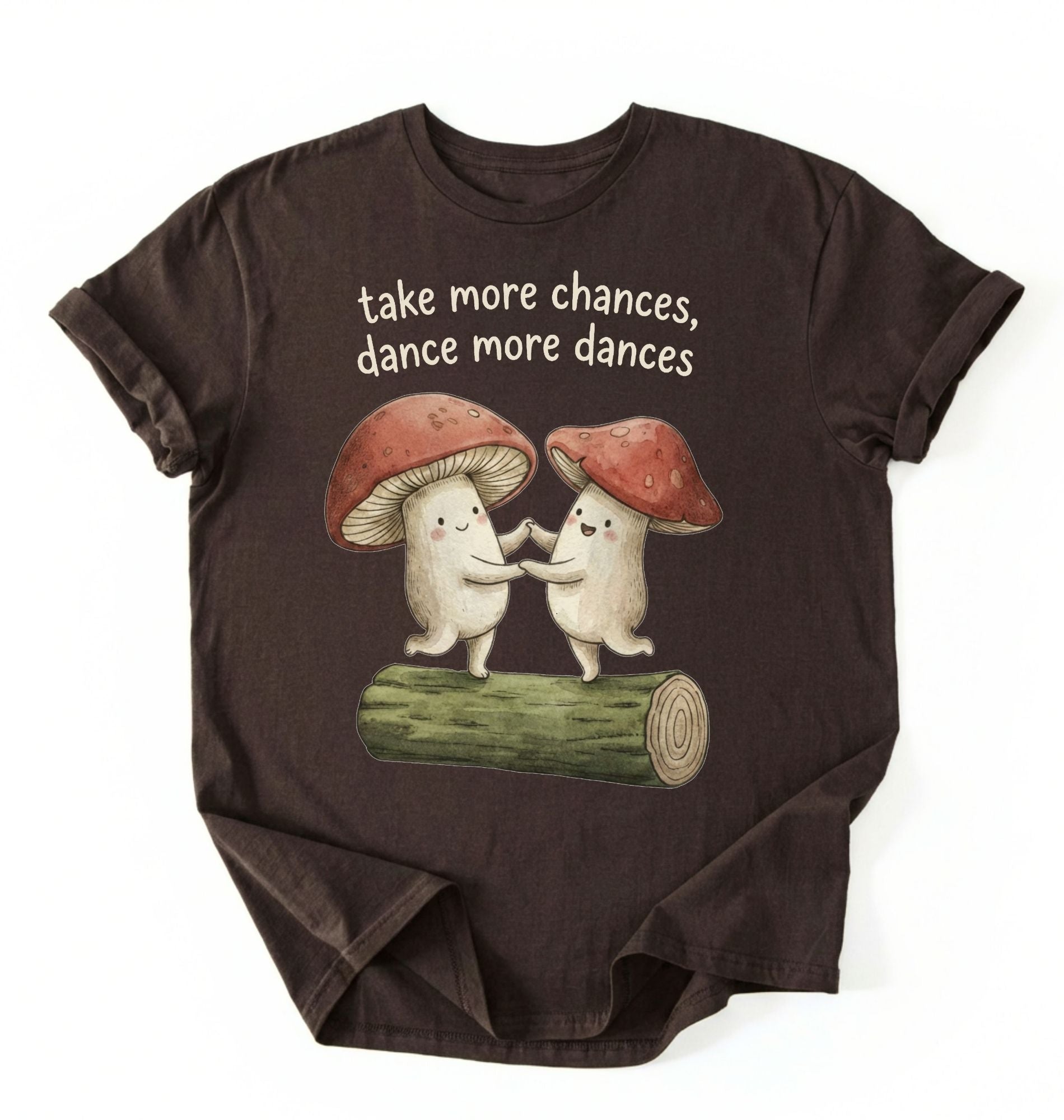 Flat lay of a dark_chocolate t-shirt with two dancing mushrooms and the phrase take more chances dance more dances, whimsical cottagecore gift tee on a white background 
