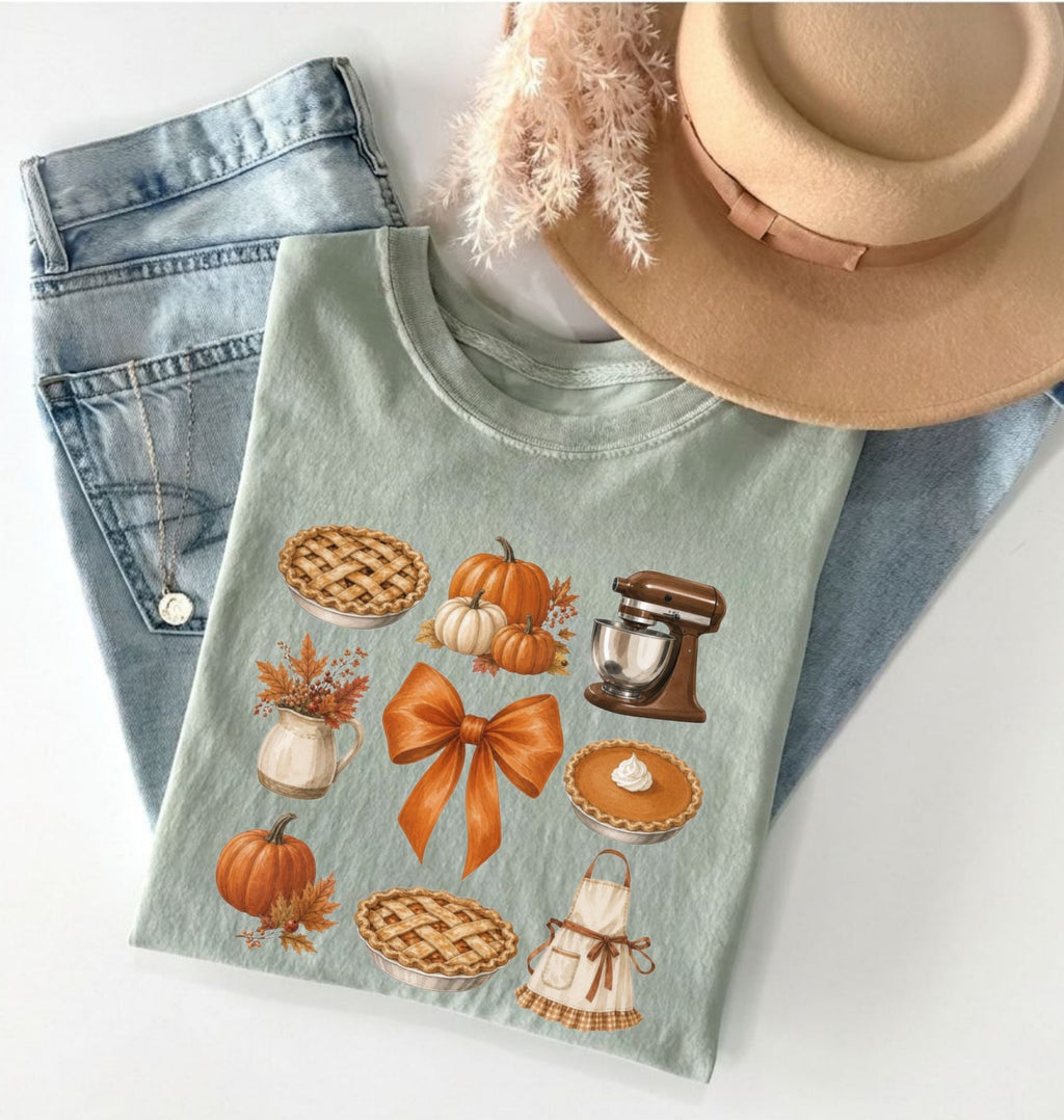 A folded sage green graphic t-shirt featuring autumn-themed illustrations of pies, pumpkins, a mixer, and a bow, styled in a flat lay with light-wash jeans, a tan hat, and dried grass on a white background.