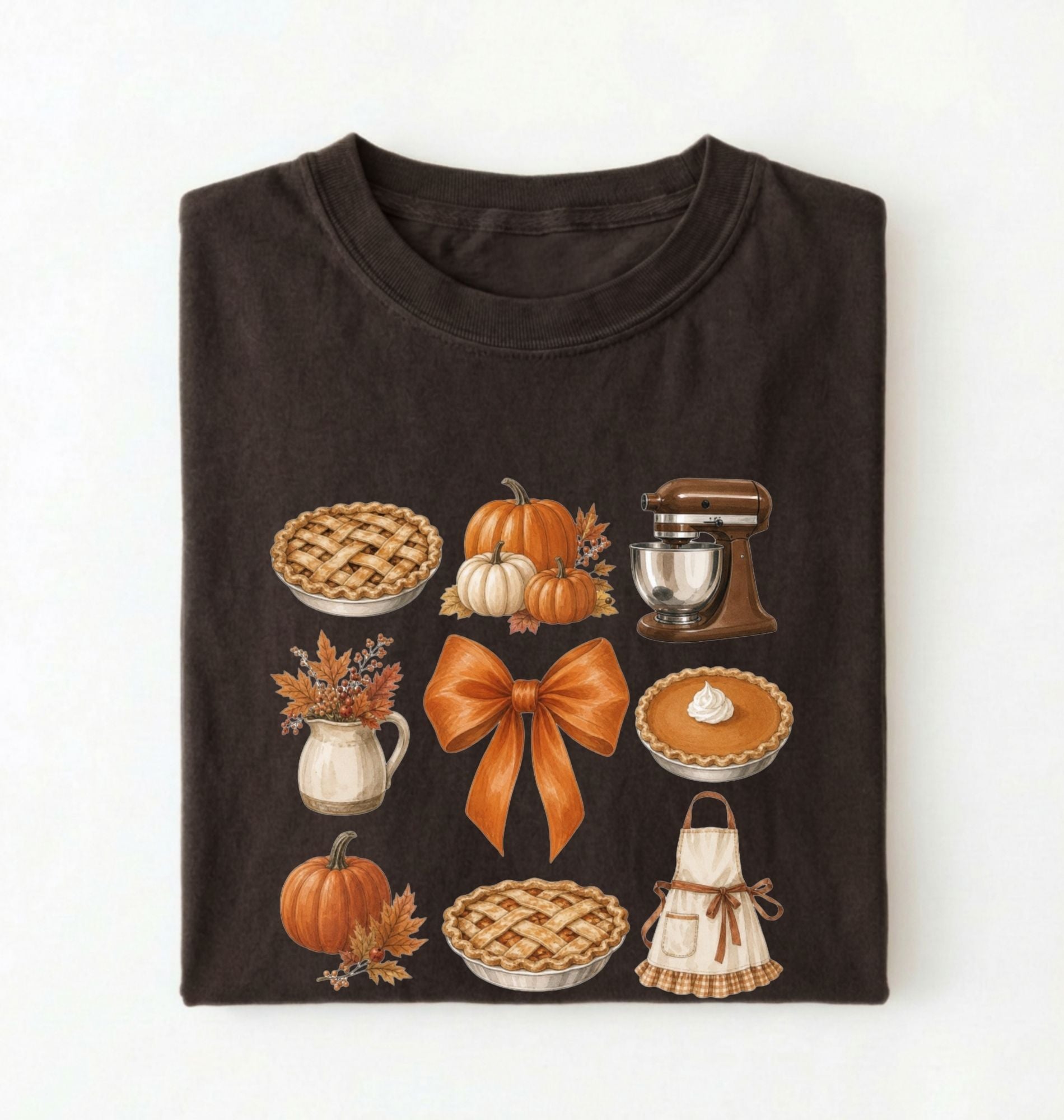 A folded dark chocolate crewneck t-shirt with a graphic print of autumn and fall baking elements, including pies, pumpkins, a stand mixer, a bow, and an apron, on a white background.