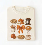 An ivory folded t-shirt featuring a watercolor-style autumn graphic print with pumpkins, pies, a stand mixer, a bow, and an apron on w white background 