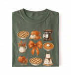 A folded dark green t-shirt with a detailed autumn-themed graphic print featuring pies, pumpkins, a stand mixer, and a bow on a white background 