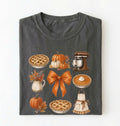 A dark gray folded t-shirt featuring a colorful autumn-themed graphic print with pumpkins, pies, a stand mixer, a bow, and an apron, displayed against a clean white background.