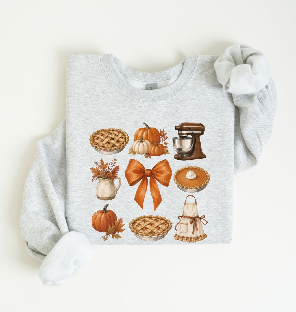 Flat lay of an ash fall baking sweatshirt featuring pumpkin pies, pumpkins, autumn leaves, a stand mixer, baking apron and orange bow on a white background 
