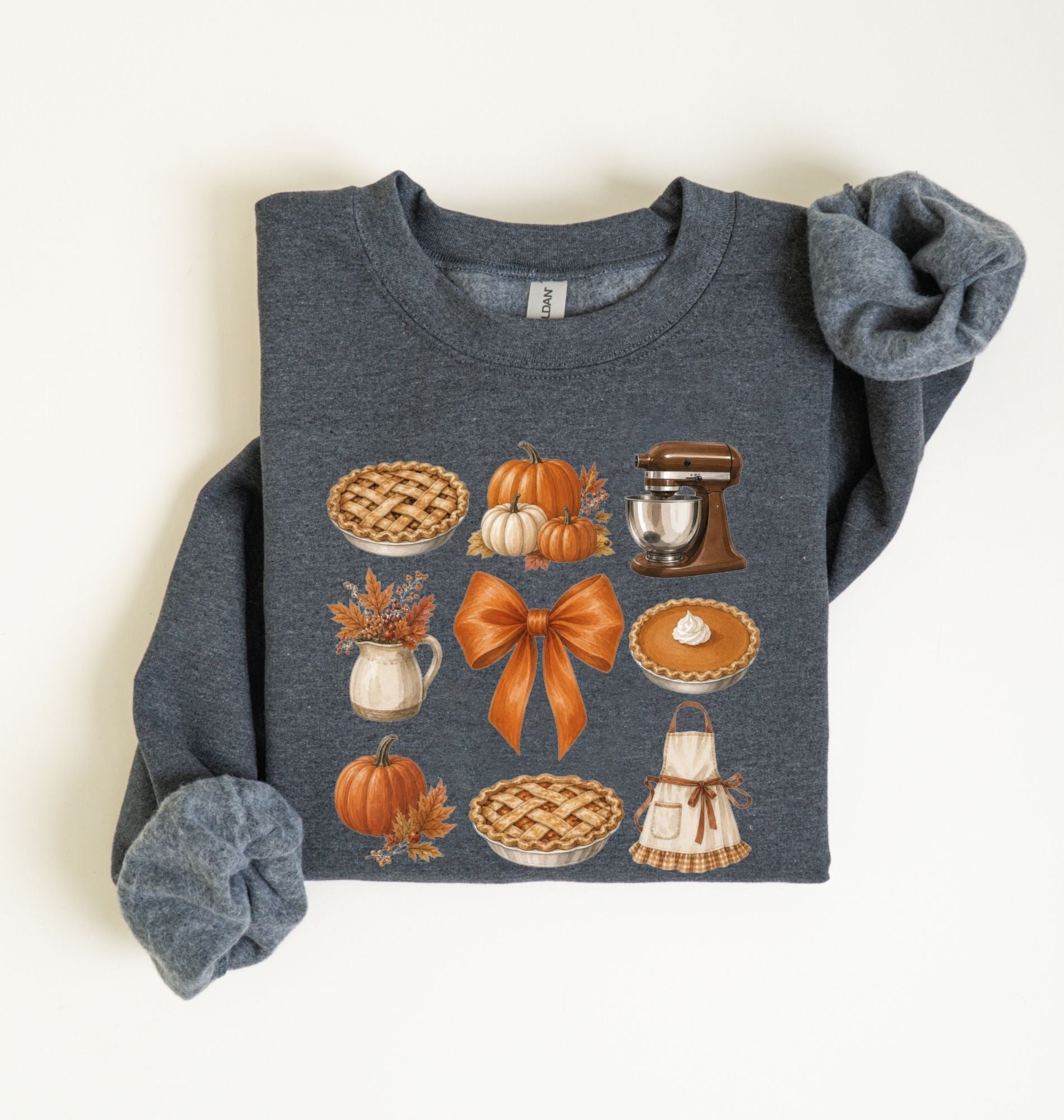 Flat lay of a dark heather fall baking sweatshirt featuring pumpkin pies, pumpkins, autumn leaves, a stand mixer, baking apron and orange bow on a white background 