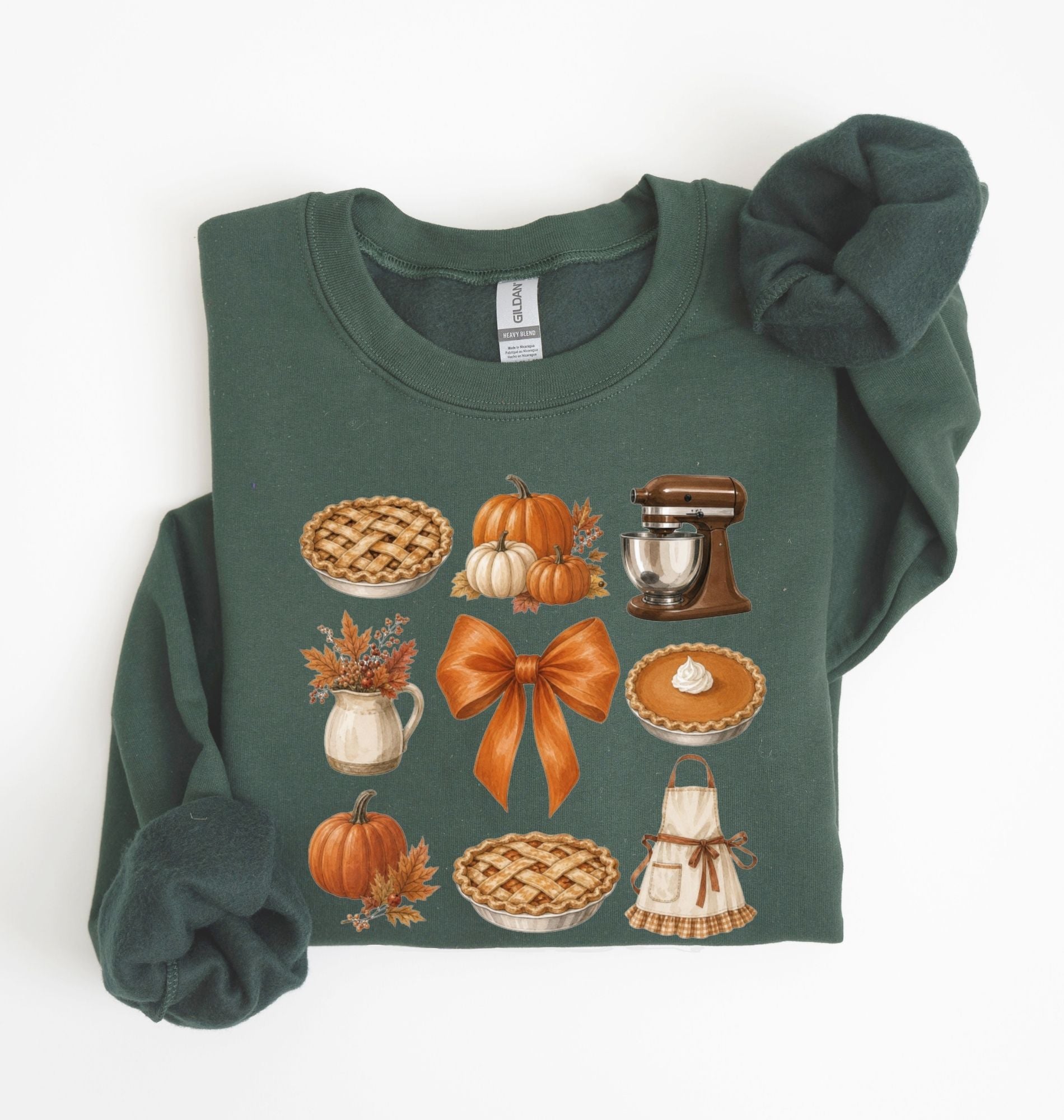 A dark green crewneck sweatshirt with long sleeves, featuring a graphic print of autumn-themed illustrations including pies, pumpkins, a stand mixer, and an orange bow. The sweatshirt is styled flat on a white background with the sleeves folded.