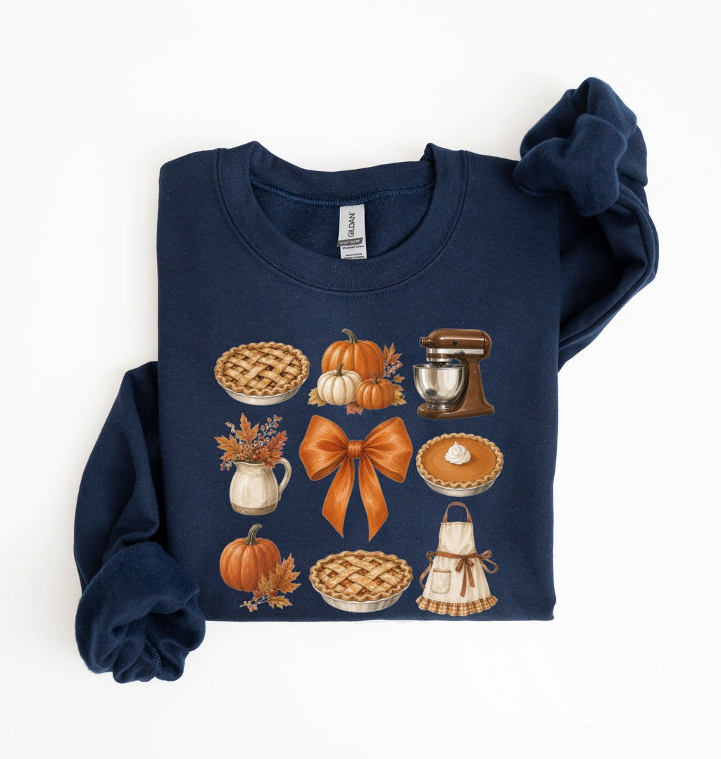 A navy blue long-sleeve crewneck sweatshirt with a festive autumn graphic print, laid flat on a white background. The graphic features illustrations of pies, pumpkins, a large orange bow, a stand mixer, and a small pumpkin.
