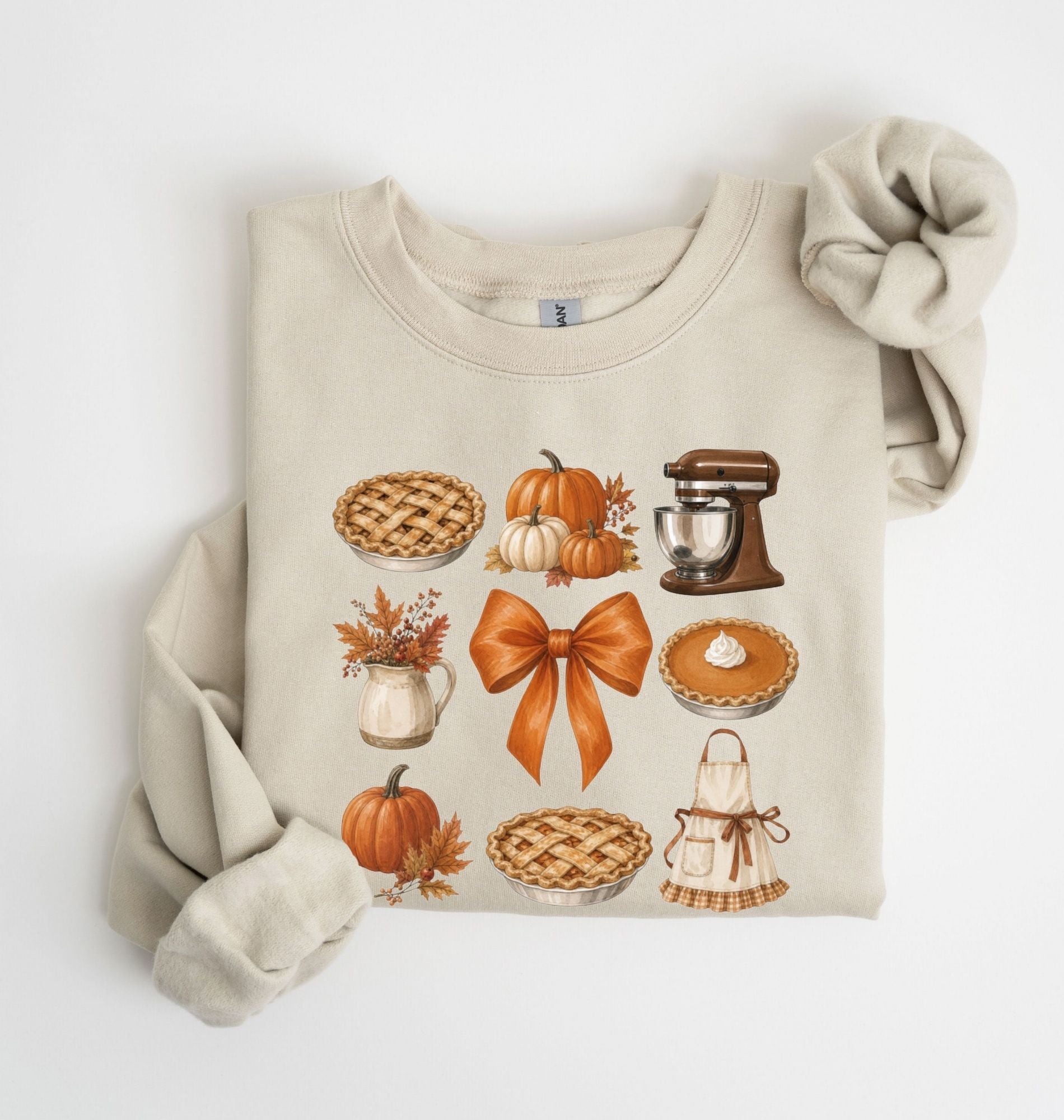 A folded beige crewneck sweatshirt with a long sleeve draped off the side, featuring a colorful autumn-themed graphic print of pies, pumpkins, a stand mixer, a pitcher of fall leaves, and an apron with a large orange bow on a white background 