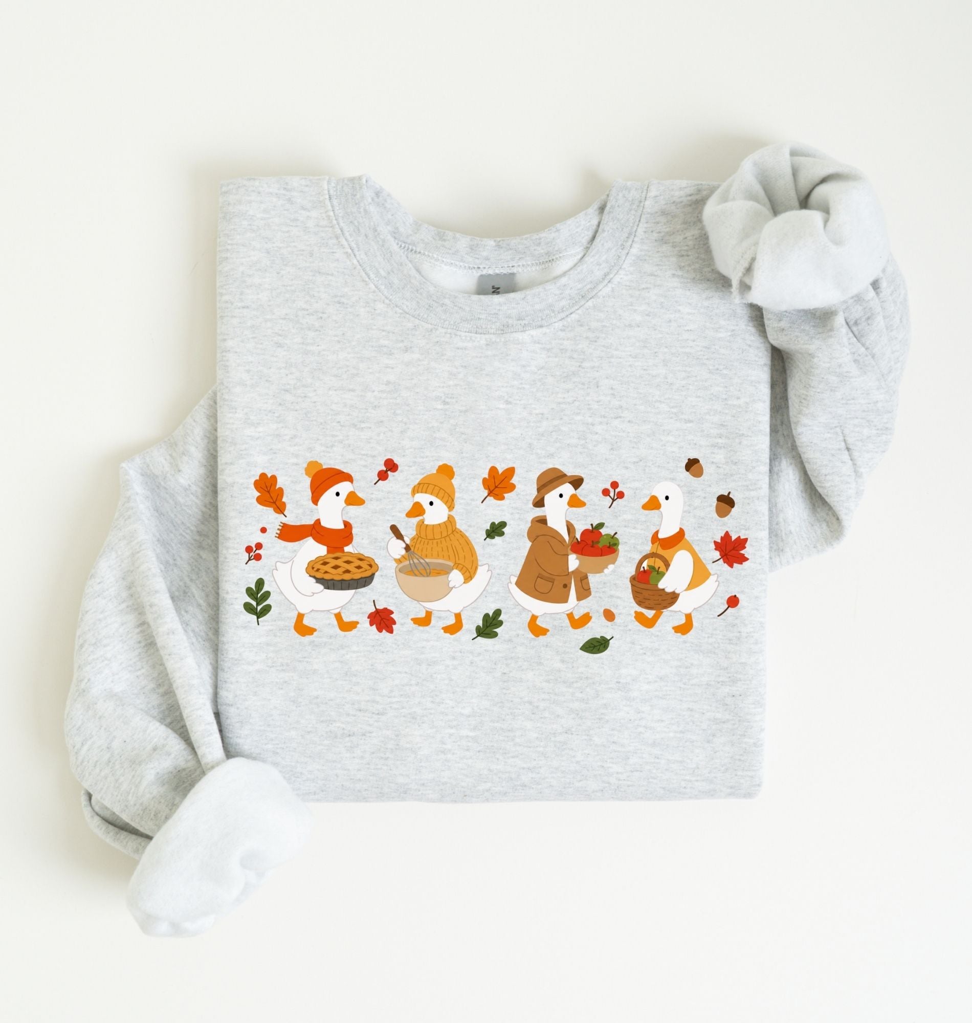 Flat lay of an ash fall sweatshirt featuring four geese baking pie, mixing ingredients and gathering apples on a white background