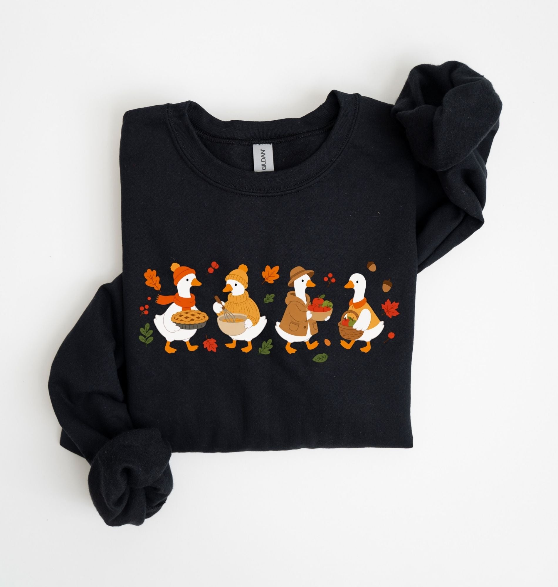 Flat lay of a white fall sweatshirt featuring four geese baking pie, mixing ingredients and gathering apples on a white background