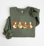 Flat lay of a military green fall sweatshirt featuring four geese baking pie, mixing ingredients and gathering apples on a white background