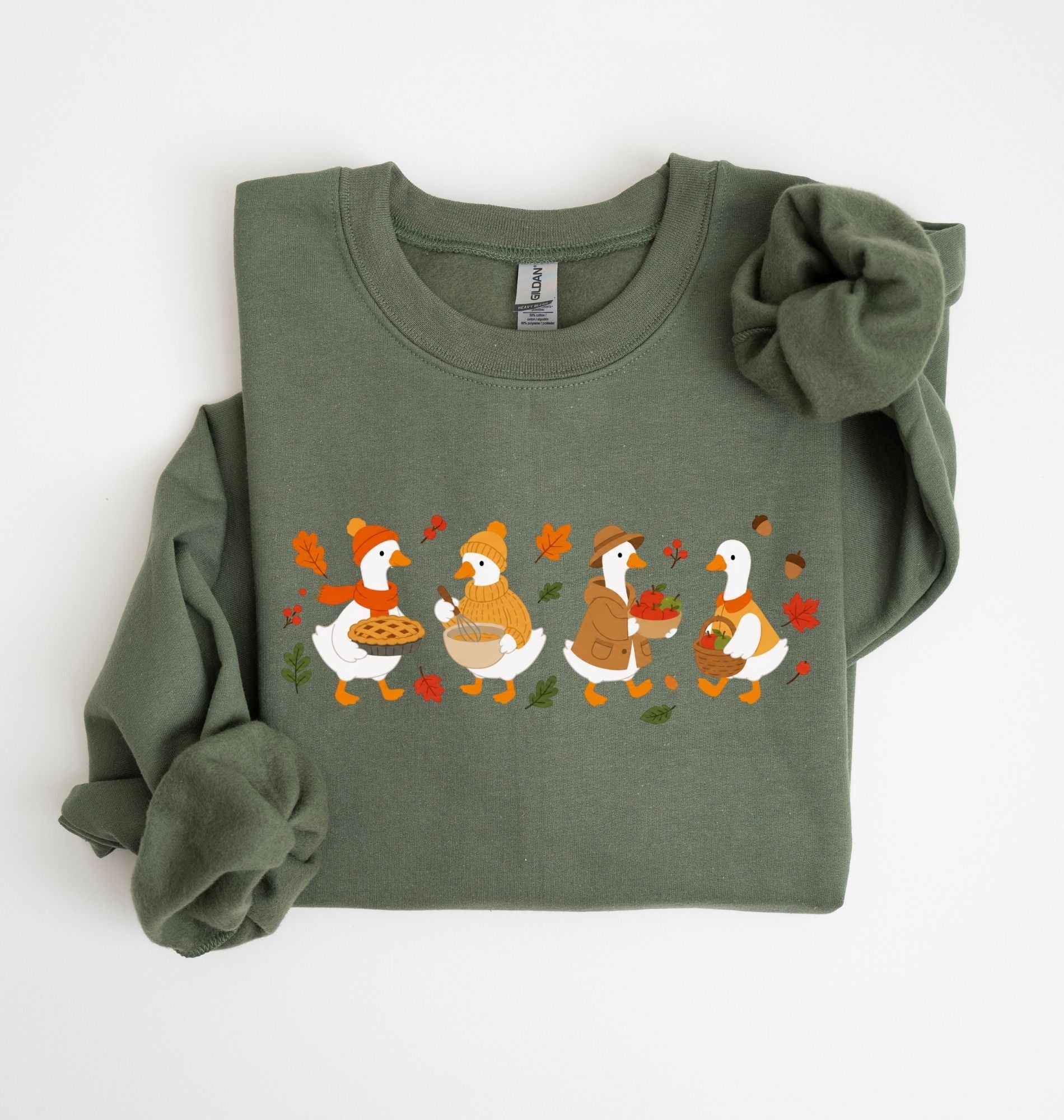 Flat lay of a military green fall sweatshirt featuring four geese baking pie, mixing ingredients and gathering apples on a white background