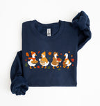 Flat lay of a blue fall sweatshirt featuring four geese baking pie, mixing ingredients and gathering apples on a white background