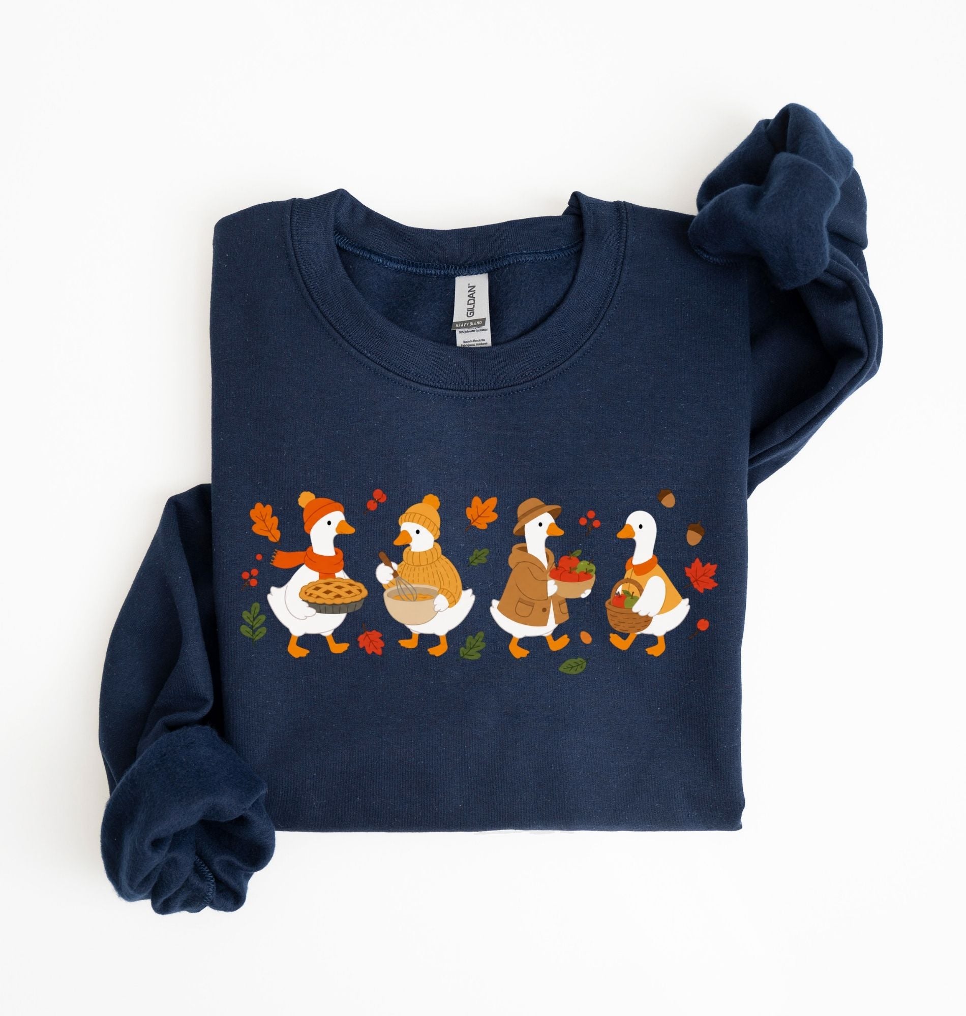 Flat lay of a blue fall sweatshirt featuring four geese baking pie, mixing ingredients and gathering apples on a white background